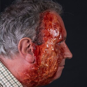May include: A side profile of a person's face with a severe, realistic wound makeup. The makeup features extensive red and brown tones, simulating a deep laceration and exposed tissue. The person has gray hair and is wearing a plaid shirt.