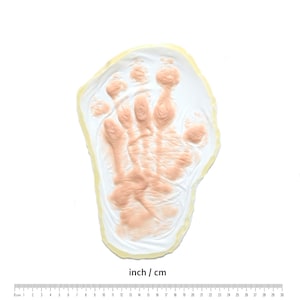 May include: A white plaster handprint mold with a light pink impression of a hand. The mold is approximately 6 inches in diameter.
