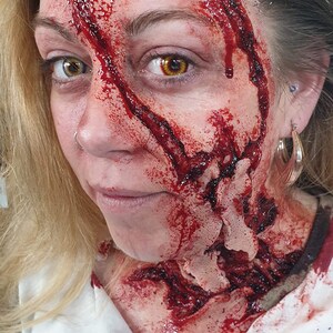 以下が含まれることがあります： 金髪の女性は顔と首に血まみれの傷を負っています。傷は深く、切り傷のようです。女性は白いシャツを着ており、服に血が付いています。