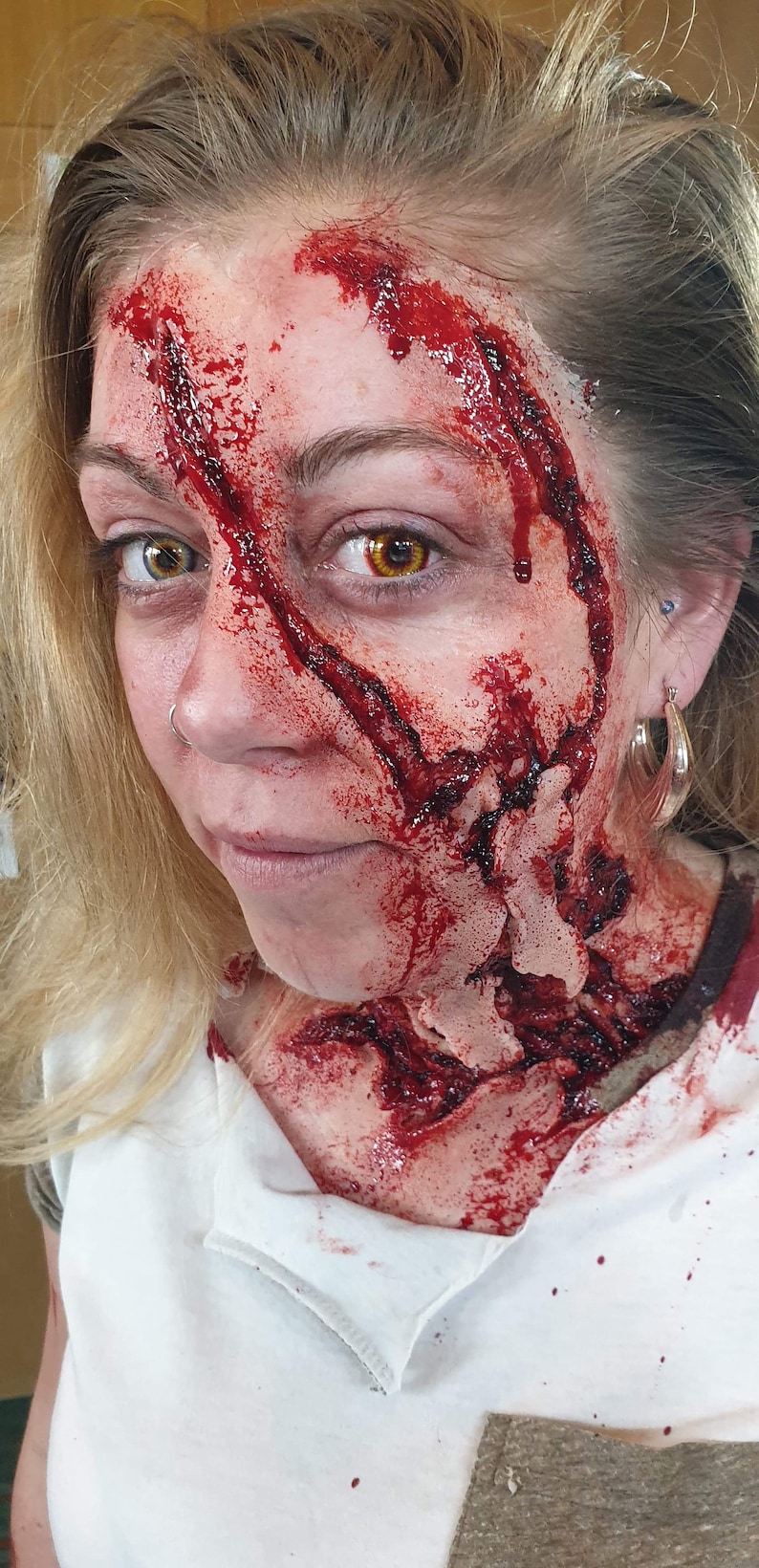 以下が含まれることがあります： 金髪の女性は顔と首に血まみれの傷を負っています。傷は深く、切り傷のようです。女性は白いシャツを着ており、服に血が付いています。
