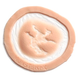 May include: A flesh-toned prosthetic wound with a circular design. The centre features a textured, open wound effect, surrounded by a raised, skin-like border. The text "MONSTERFX" is embossed on the border.