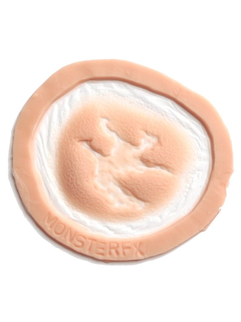 May include: A flesh-toned prosthetic wound with a circular design. The centre features a textured, open wound effect, surrounded by a raised, skin-like border. The text "MONSTERFX" is embossed on the border.