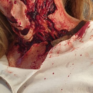 以下が含まれることがあります： 女性の首のクローズアップで、大きな血まみれの傷があります。傷は開いていて生々しく、血が首を伝って白いシャツに滴っています。女性は金のイヤリングをしています。