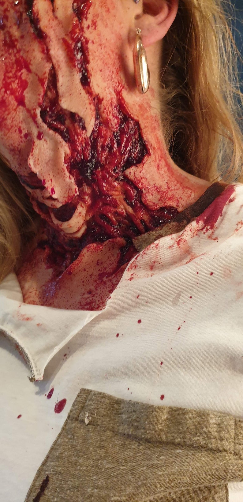 以下が含まれることがあります： 女性の首のクローズアップで、大きな血まみれの傷があります。傷は開いていて生々しく、血が首を伝って白いシャツに滴っています。女性は金のイヤリングをしています。