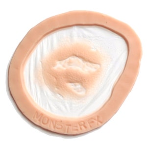 Puede incluir: Primer plano de una pr&oacute;tesis de cicatriz de silicona de color carne con una superficie texturizada. La cicatriz est&aacute; rodeada por un borde rosa claro. El texto "MONSTERFX" est&aacute; impreso en el borde.