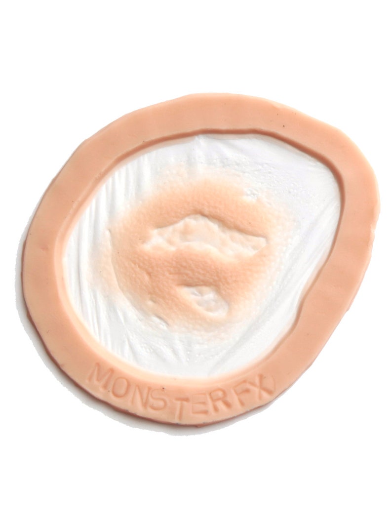 Puede incluir: Primer plano de una pr&oacute;tesis de cicatriz de silicona de color carne con una superficie texturizada. La cicatriz est&aacute; rodeada por un borde rosa claro. El texto "MONSTERFX" est&aacute; impreso en el borde.