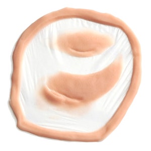 Puede incluir: Un accesorio de herida prot&eacute;sica de color carne. El accesorio tiene forma circular con un borde elevado y detalles interiores, dise&ntilde;ado para simular una lesi&oacute;n cut&aacute;nea. El producto est&aacute; hecho de un material flexible y est&aacute; sobre un fondo blanco.