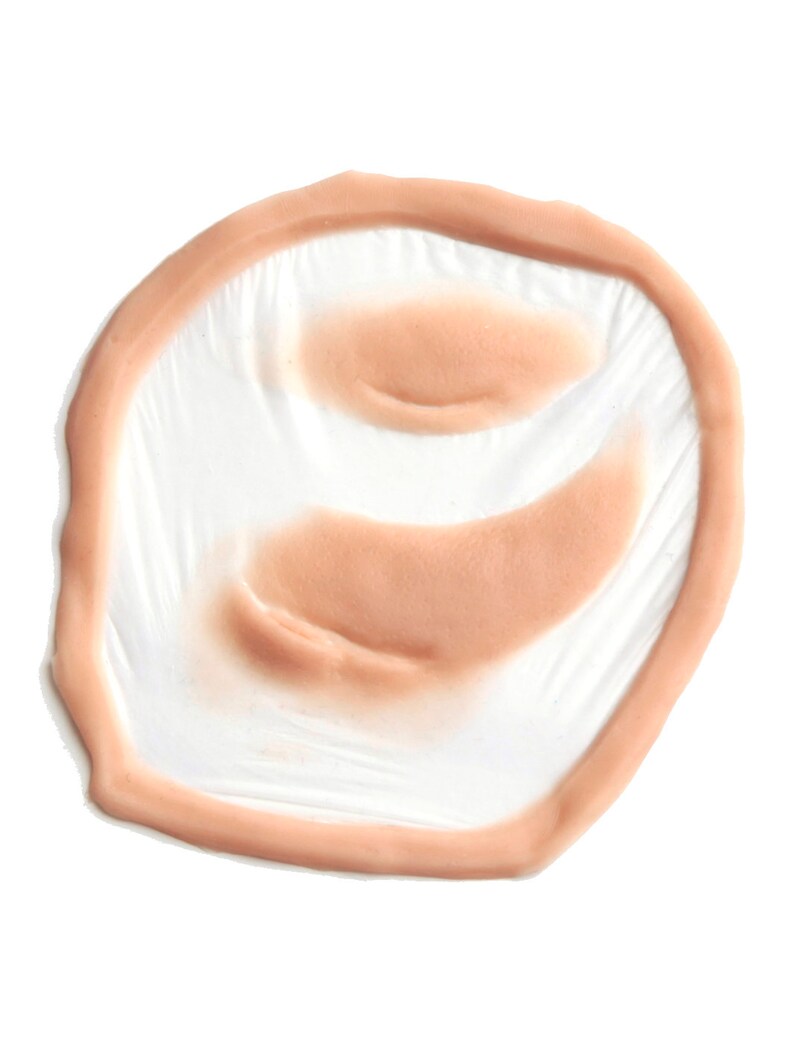 Puede incluir: Un accesorio de herida prot&eacute;sica de color carne. El accesorio tiene forma circular con un borde elevado y detalles interiores, dise&ntilde;ado para simular una lesi&oacute;n cut&aacute;nea. El producto est&aacute; hecho de un material flexible y est&aacute; sobre un fondo blanco.