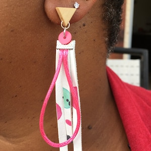 May include: A pair of dangle earrings with a pink and white leather tassel and a wooden triangle charm. The earrings are pink and white with a geometric pattern.