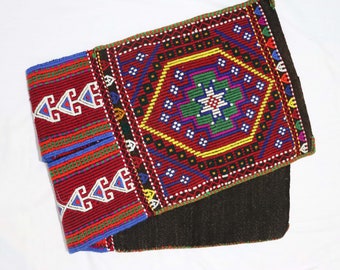 May include: Handmade textile with a geometric design. The main color is a deep red, accented with blue, green, yellow, and white. The design features repeating patterns and shapes, with a dark brown backing.