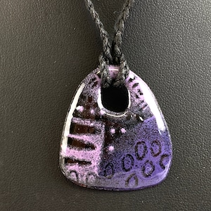 May include: A purple and black ceramic pendant with a geometric design. The pendant is attached to a black cord.