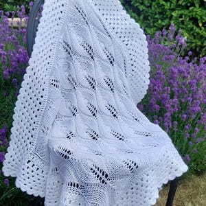 May include: A white, crocheted blanket with a scalloped edge is draped over a black metal chair. The blanket features a detailed openwork design with a repeating pattern of diamond shapes. The background includes green foliage and purple flowers.