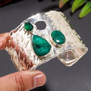 May include: A wide silver cuff bracelet featuring a textured, hammered surface with three bezel-set green gemstones. The stones include one small pear-shaped gem, one large pear-shaped gem, and one oval-shaped gem, all displaying deep green hues.