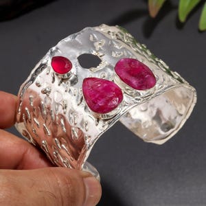 Puede incluir: Un brazalete ancho de plata con una superficie martillada y texturizada, adornado con tres piedras preciosas de rubí rosa intenso en forma de lágrima engastadas en bisel. El metal tiene un acabado pulido y brillante con irregularidades.