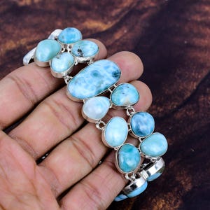 Op de afbeelding: Een zilveren armband met meerdere ovale en afgeronde larimar stenen in tinten lichtblauw en turquoise. De stenen zijn gezet in individuele zilveren bezels en met elkaar verbonden. De armband wordt gepresenteerd op een bruine achtergrond.