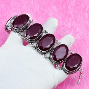 May include: A bracelet featuring five large, oval-shaped, faceted deep red gemstones set in ornate silver bezels. The stones are linked together by small silver rings, creating a flexible chain design with a metallic finish and intricate detailing.