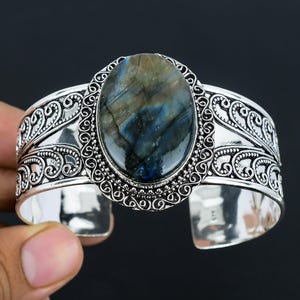 Puede incluir: Pulsera de brazalete de plata con una piedra preciosa de labradorita de forma ovalada. La pulsera tiene un diseño detallado y adornado con espirales y volutas.