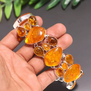 Peut inclure: Un bracelet en argent orné de multiples pierres d'ambre de formes et de tailles variées. Les pierres d'ambre varient du jaune clair à l'orange foncé. Le bracelet est tenu dans une main sur fond sombre.
