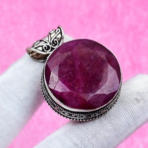 May include: A large, faceted ruby gemstone pendant with a silver-coloured bail. The ruby is a deep, rich red colour and is set in a detailed, ornate silver setting. The pendant is displayed against a pink background.