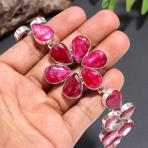 Puede incluir: Una pulsera con múltiples piedras preciosas de rubí rosa intenso, facetadas y en forma de lágrima, engastadas en metal plateado pulido. El diseño central consta de seis piedras dispuestas en forma de flor, unidas por eslabones de plata.