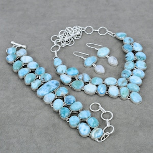 May include: A jewelry set featuring a bracelet, necklace, and earrings. The set is made of silver and features oval-shaped, light blue gemstones. The earrings have a teardrop-shaped, white gemstone. The jewelry is displayed on a gray surface.