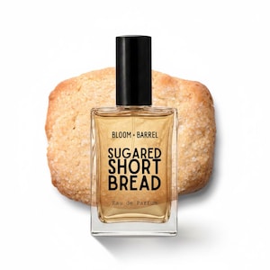 May include: A clear glass perfume bottle with a black cap containing golden liquid. The label reads BLOOM + BARREL SUGARED SHORT BREAD Eau de Parfum. The bottle is positioned in front of a round, golden-brown cookie with a visible sugar-coated texture.