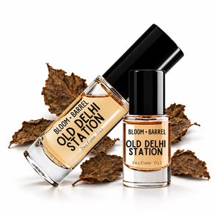 May include: Two clear glass bottles of Bloom + Barrel Old Delhi Station perfume oil with black caps. The larger bottle is positioned diagonally behind the smaller upright bottle. Both contain amber-colored liquid and feature black text labels.