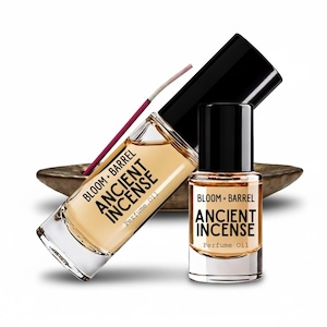 May include: Two clear glass bottles of Bloom + Barrel Ancient Incense perfume oil with black caps. The larger bottle is angled, and the smaller bottle stands upright. A single thin purple incense stick rests behind the bottles.