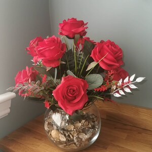 May include: A glass vase filled with red roses and decorative greenery. The vase is filled with small, brown stones.