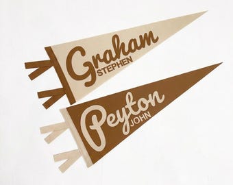 May include: Two triangular felt pennants with ties at the base. The top pennant is cream with brown text reading Graham STEPHEN. The bottom pennant is brown with cream text reading Peyton JOHN. Both feature a large script name and smaller block text.
