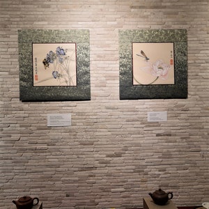 K&ouml;nnte beinhalten: Zwei gerahmte asiatische Bilder an einer wei&szlig;en Backsteinwand. Das Bild links zeigt eine Biene auf einer blauen Blume. Das Bild rechts zeigt eine Libelle auf einer rosa Lotusblume. Unter den Bildern befinden sich drei Teetassen und eine Teekanne auf einem Holztablett.