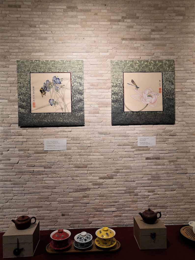 K&ouml;nnte beinhalten: Zwei gerahmte asiatische Bilder an einer wei&szlig;en Backsteinwand. Das Bild links zeigt eine Biene auf einer blauen Blume. Das Bild rechts zeigt eine Libelle auf einer rosa Lotusblume. Unter den Bildern befinden sich drei Teetassen und eine Teekanne auf einem Holztablett.