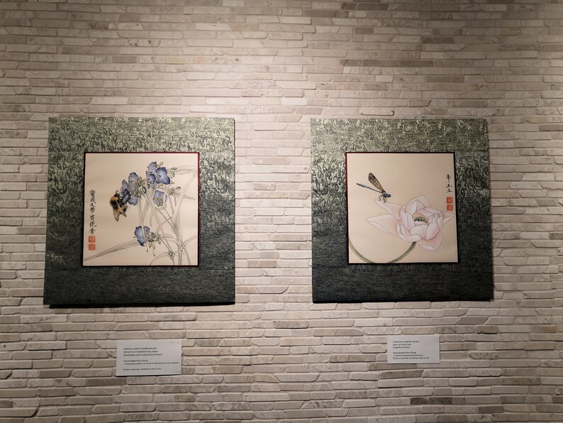 K&ouml;nnte beinhalten: Zwei gerahmte asiatische Gem&auml;lde an einer wei&szlig;en Backsteinwand. Das linke Gem&auml;lde zeigt eine Biene auf einer Blume mit chinesischen Schriftzeichen. Das rechte Gem&auml;lde zeigt eine Libelle auf einer Lotusblume mit chinesischen Schriftzeichen.
