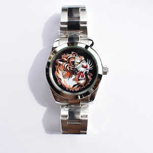 Puede incluir: Un reloj de pulsera plateado con una esfera negra y un gráfico de tigre. El reloj tiene una banda de metal plateado.