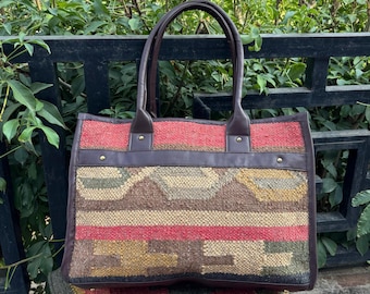 May include: A rectangular tote bag with a woven, patterned front in shades of red, brown, and yellow. The bag has brown leather handles and trim, with brass-colored rivets. The bag is sitting on a patterned pillow.
