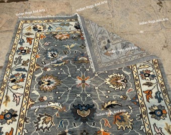 Puede incluir: Una alfombra gris y blanca con un patrón floral. La alfombra tiene un borde con un patrón repetitivo de flores y hojas. La alfombra está doblada por la mitad, mostrando el reverso de la alfombra.