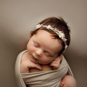 Könnte beinhalten: Ein neugeborenes Baby-Mädchen schläft friedlich, eingewickelt in ein beiges Swaddle. Sie trägt ein weißes Blumenkronen-Haarband.