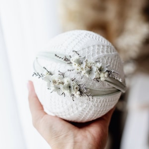 May include: A white lace headband with a delicate floral design and a light green ribbon. The headband is held in a hand.