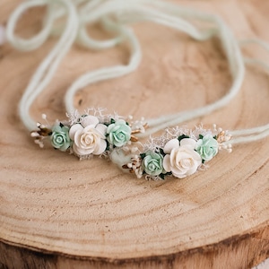 Puede incluir: Un par de diademas florales delicadas con flores de tela blancas y verdes. Las diademas están hechas de un hilo verde claro.