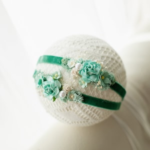 Op de afbeelding: Een groen fluwelen hoofdband met een bloemenontwerp met lichtblauwe en witte bloemen. Het hoofdband is op een witte achtergrond.
