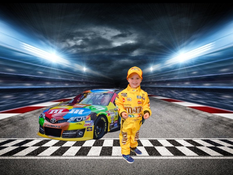 Op de afbeelding: Een jonge jongen in een geel racepak met het M&M's-logo staat voor een raceauto met het M&M's-logo. De auto staat op een racecircuit met een geblokte finishlijn.