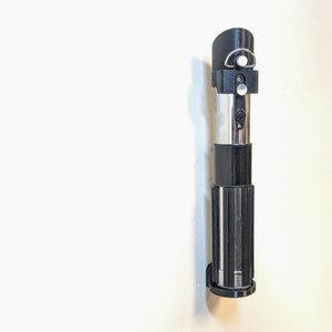 May include: A black lightsaber wall mount with a silver blade and a black handle. The mount has a small silver knob on the side.