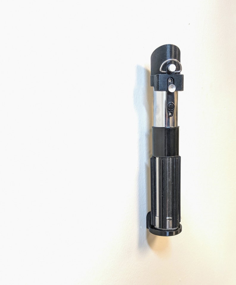 May include: A black lightsaber wall mount with a silver blade and a black handle. The mount has a small silver knob on the side.