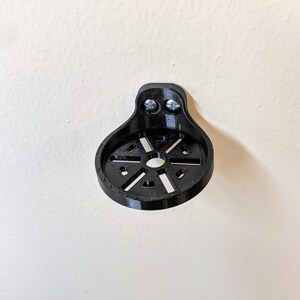 May include: A black plastic wall mount with a circular design and two screw holes. The mount is designed to hold a small object, such as a camera or a phone.