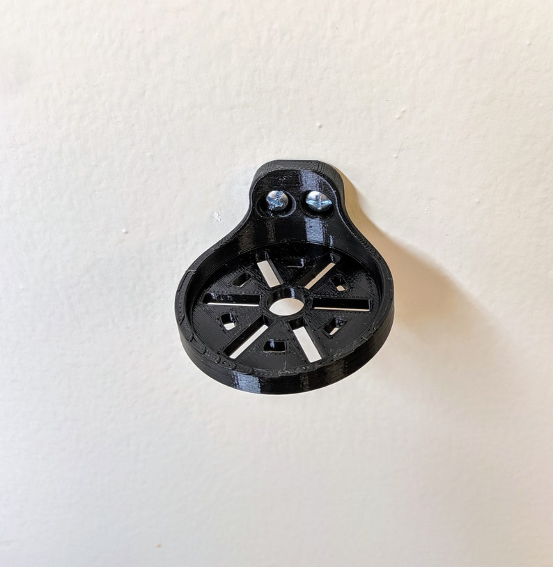 May include: A black plastic wall mount with a circular design and two screw holes. The mount is designed to hold a small object, such as a camera or a phone.