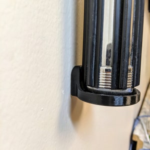 May include: A black plastic wall mount for a lightsaber. The mount is designed to hold the lightsaber securely in place.