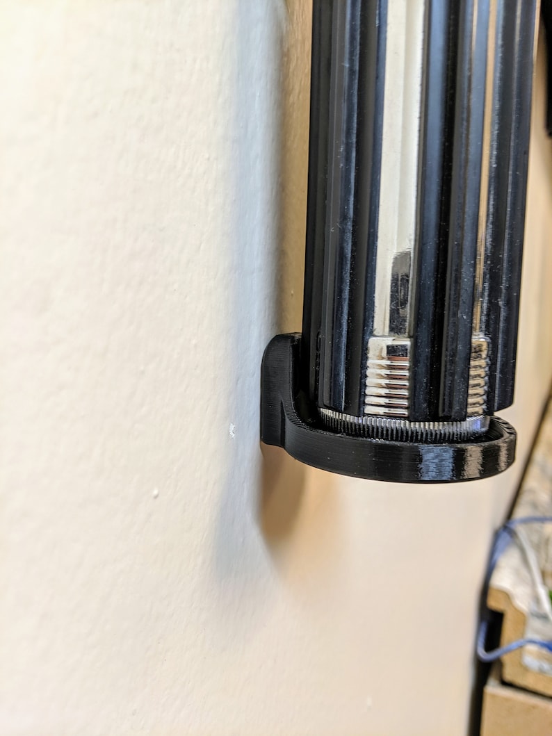May include: A black plastic wall mount for a lightsaber. The mount is designed to hold the lightsaber securely in place.