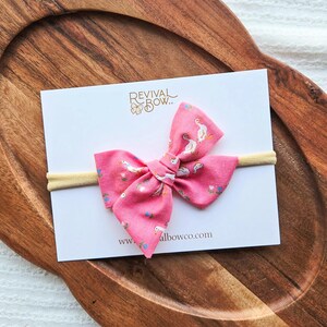 Op de afbeelding: Een roze stoffen strik met een wit ganzenpatroon, bevestigd aan een gele haarband. De strik ligt op een witte kaart met de tekst "REVIVAL BOW" en het websiteadres "www.albowco.com". De strik wordt gepresenteerd op een houten dienblad.