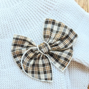 May include: A decorative hair bow featuring a brown, tan, and cream plaid pattern with a central knot, positioned on a textured cream-colored knit sweater. The fabric of the bow has a visible woven grid design and finished edges.