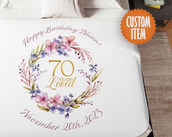 May include: White blanket with a floral wreath design. The wreath features pink and purple flowers, green leaves, and the text "Happy Birthday Nana!" and "70 Years Loved." The date "November 20th, 2023" is also included. An orange "Custom Item" tag is visible.